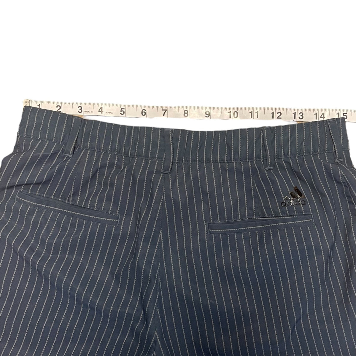 A pair of gray pinstripe shorts laid flat on a white background, with a measuring tape laid across the waistband to show the size. The Adidas logo is visible on the back pocket.
