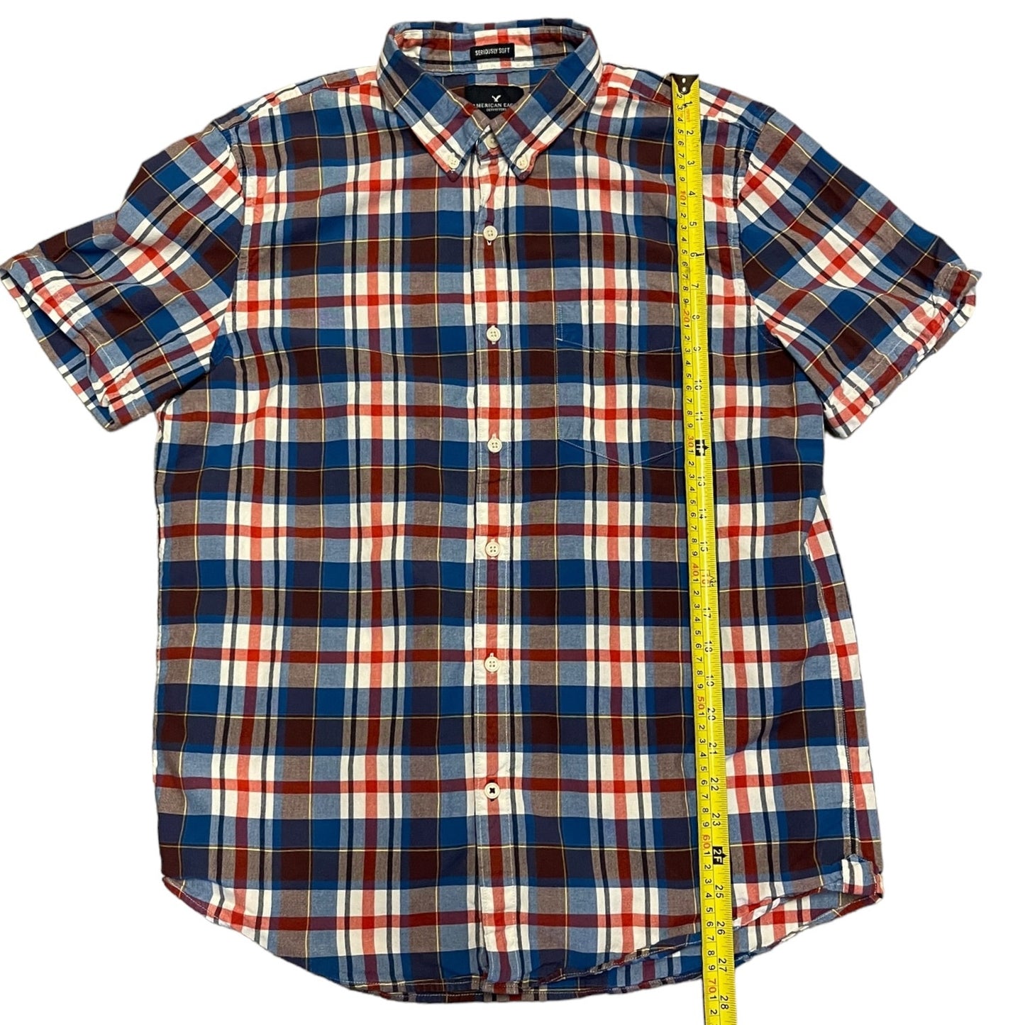 A short-sleeve button-down plaid shirt in blue, red, white, and brown, shown flat with a yellow measuring tape running vertically along the side.