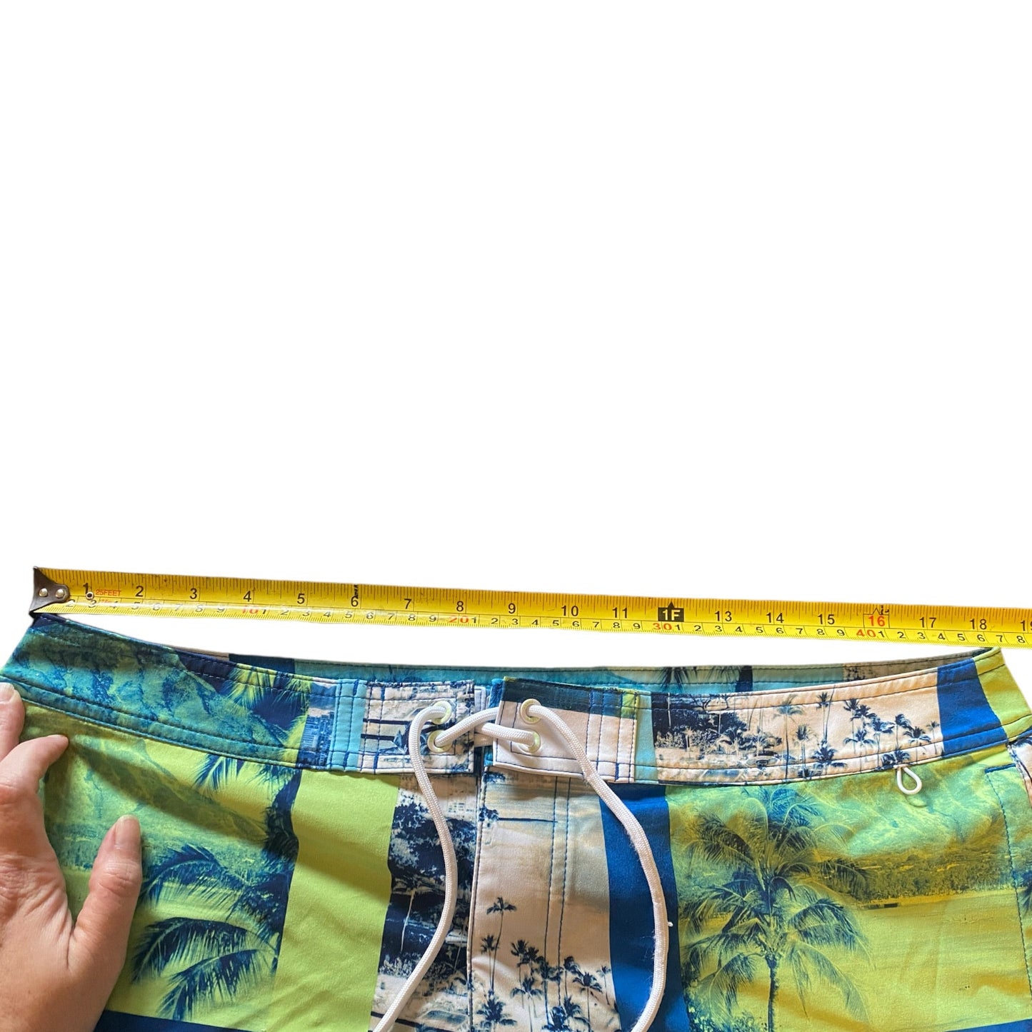 A person's hand holding down a yellow tape measure across the waistband of colorful swim trunks. The swim trunks feature a tropical print with blue palm trees, yellow-green sand, and blue denim-style panels, with a white drawstring.