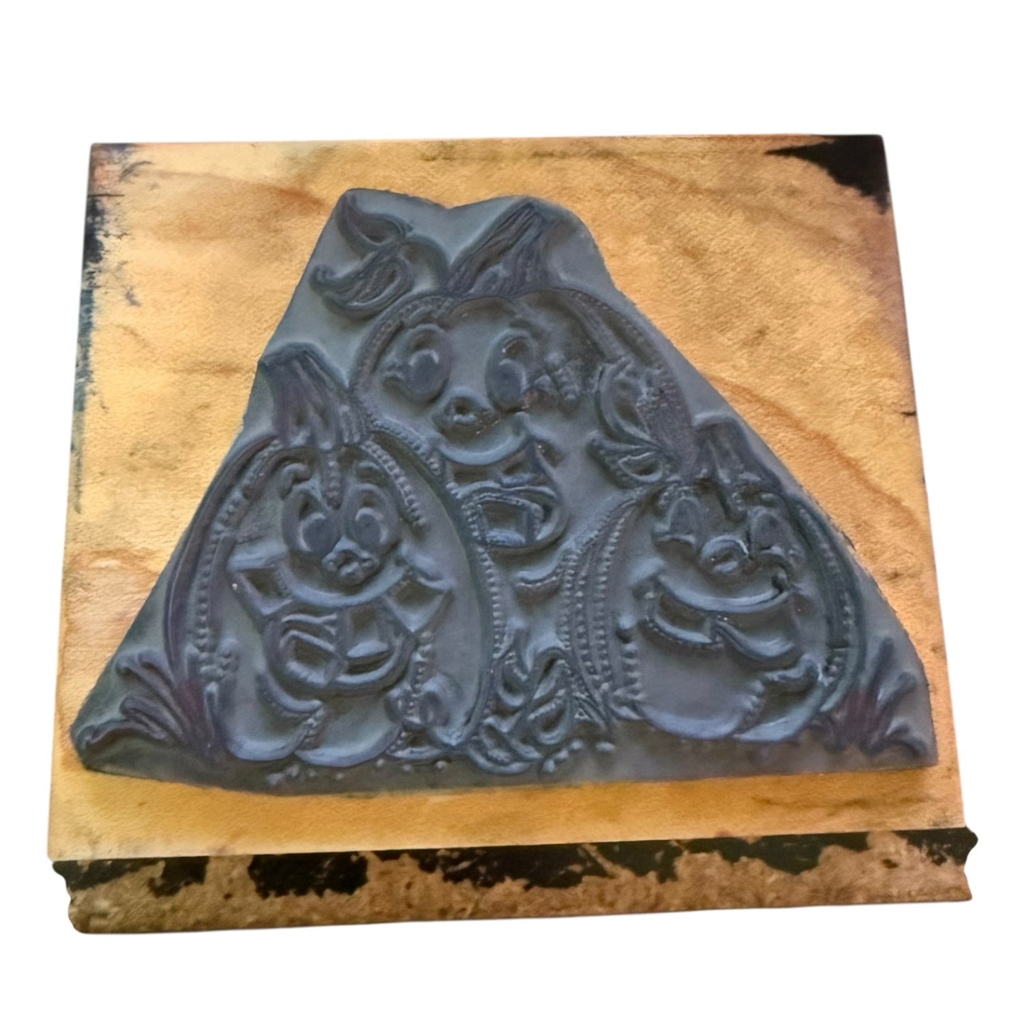 A dark gray rubber stamp with smiling pumpkins, mounted on a square tan textured board against a white background.