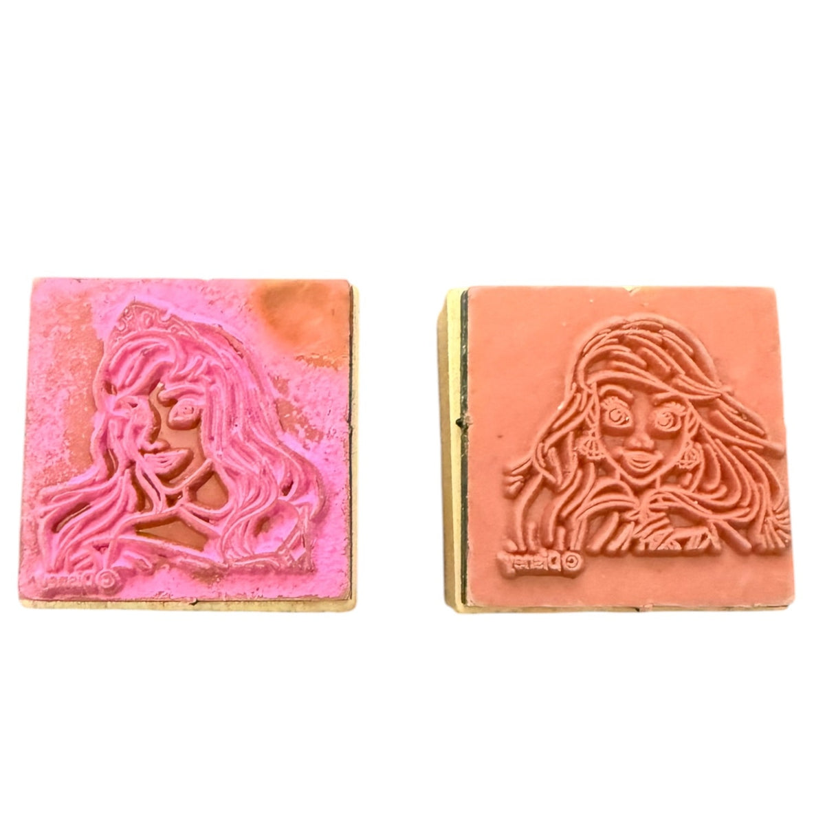 Two square wooden stamp blocks with carved female face profiles, one in pink and one in terracotta, shown side by side on a white background.
