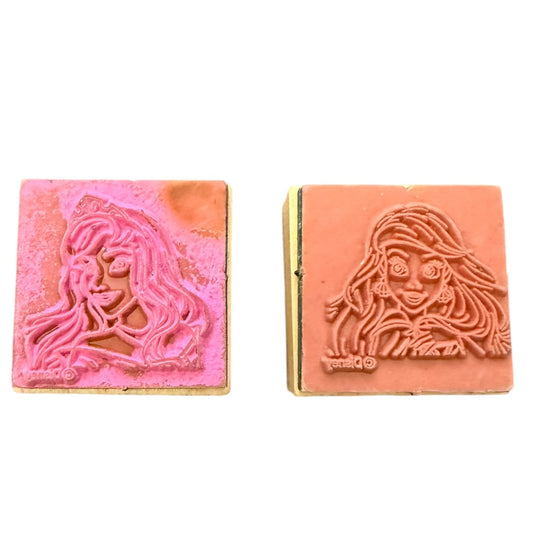 Two square wooden stamp blocks with carved female face profiles, one in pink and one in terracotta, shown side by side on a white background.