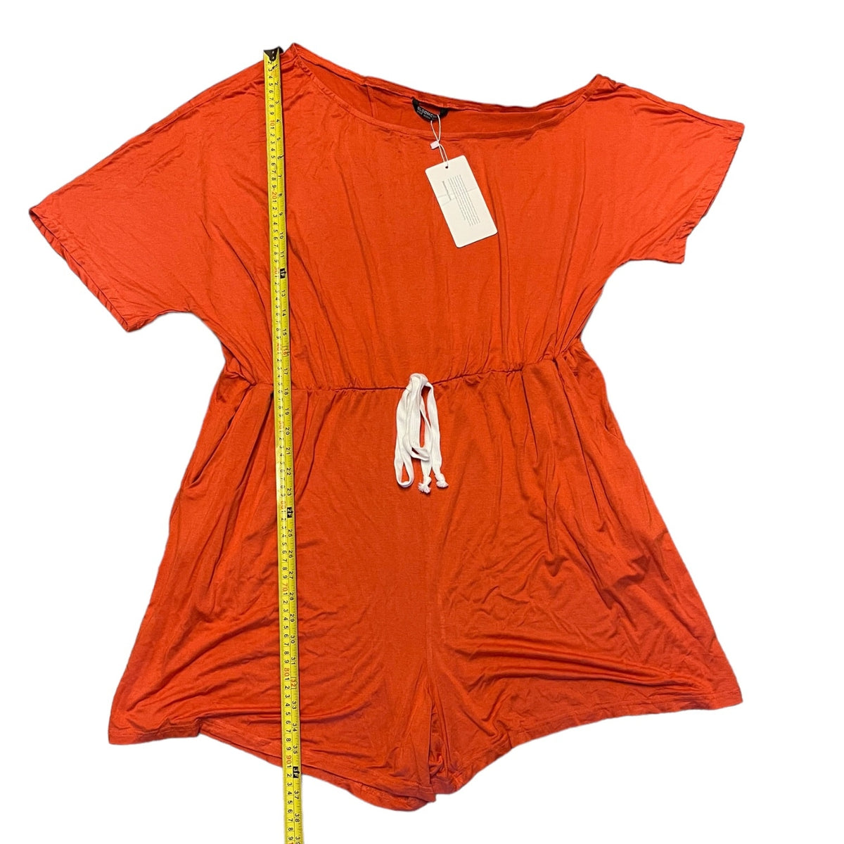 An orange short-sleeve romper , laid flat on a white background with a yellow measuring tape standing vertically on the left side.