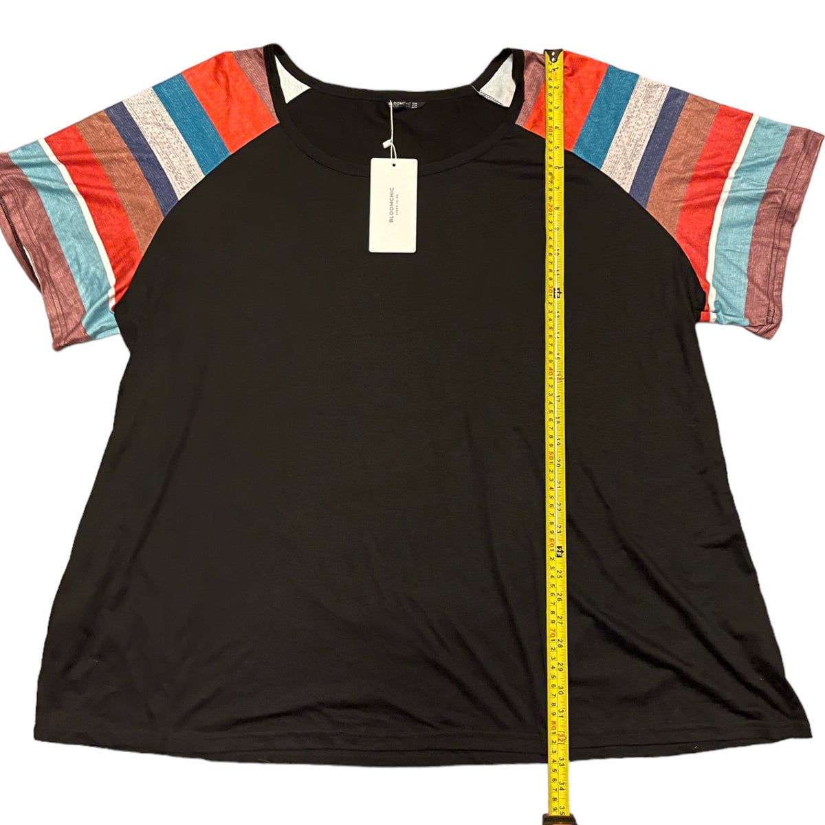 A black top with multicolor striped sleeves, shown flat-laid with a yellow measuring tape vertically aligned next to it for scale.