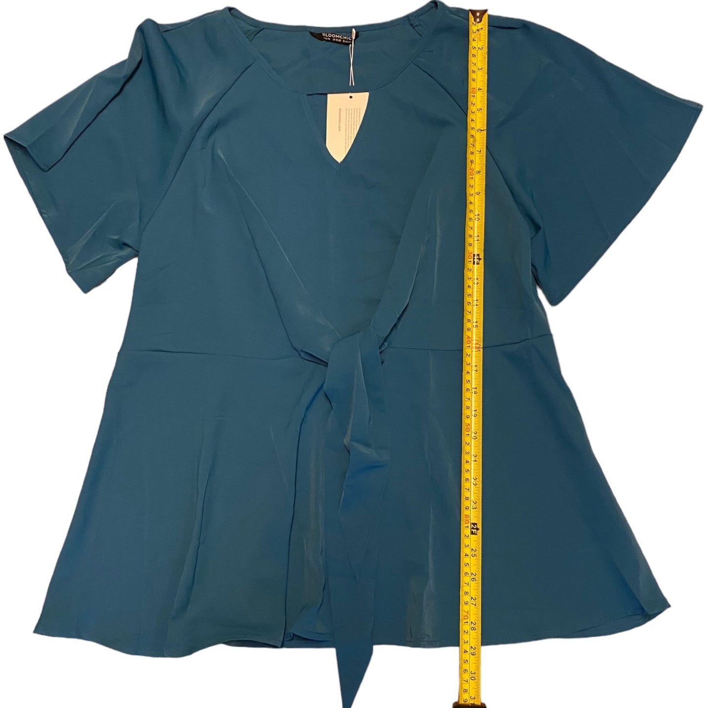 A short-sleeve wrap-style top with a tie-front detail, shown flat against a white background with a yellow measuring tape standing vertically to indicate size.