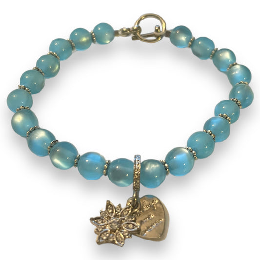 A beaded bracelet with iridescent light blue beads separated by small silver spacer rings, featuring a silver-tone toggle clasp and two dangling charms: a snowflake charm and a heart charm with engraved text, shown on a white background.