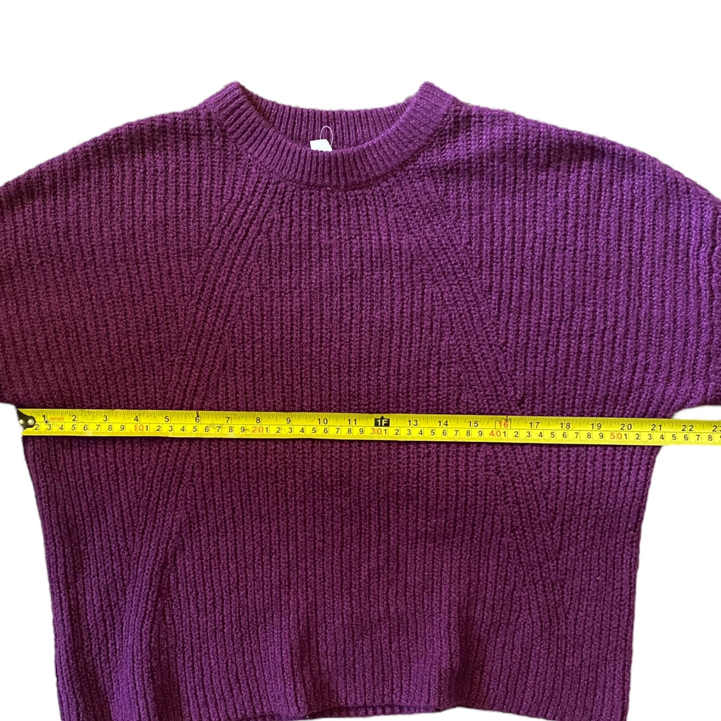 A purple ribbed knit sweater laid flat on a white background with a yellow tape measure stretched horizontally across the chest to show the width.