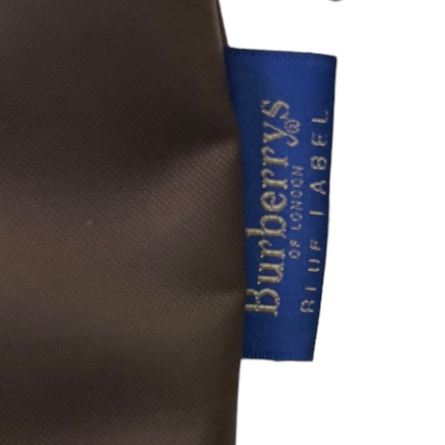 Close-up of a dark brown textured tote with a blue Burberry of London Blue Label sewn onto the side, against a white background.