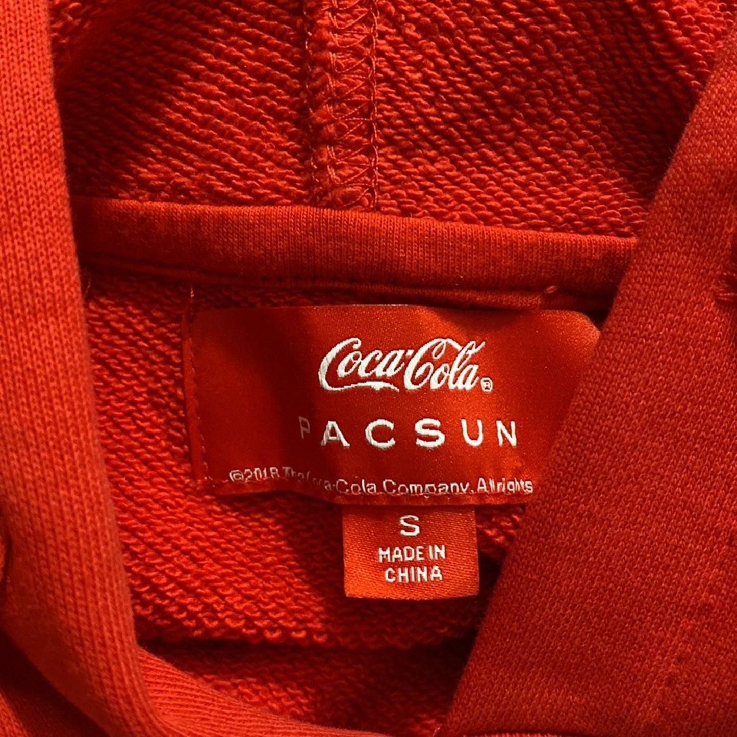 Close-up of a red knit garment showing the inner neck label with Coca-Cola and PACSUN branding, plus a small tag reading Made in China.