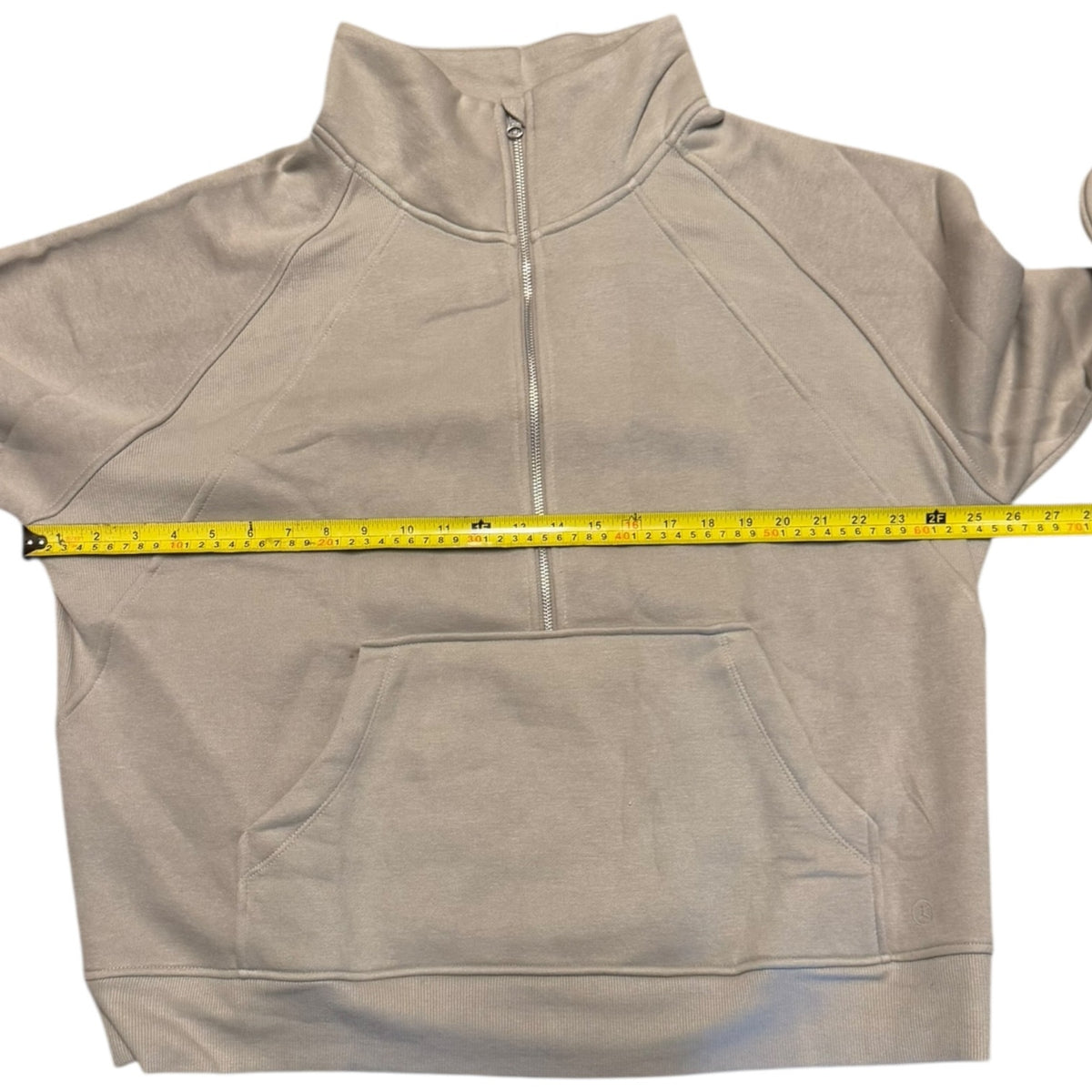 A half-zip pullover hoodie with a front kangaroo pocket, shown with a yellow tape measure stretched horizontally across the chest to indicate the garment's width.