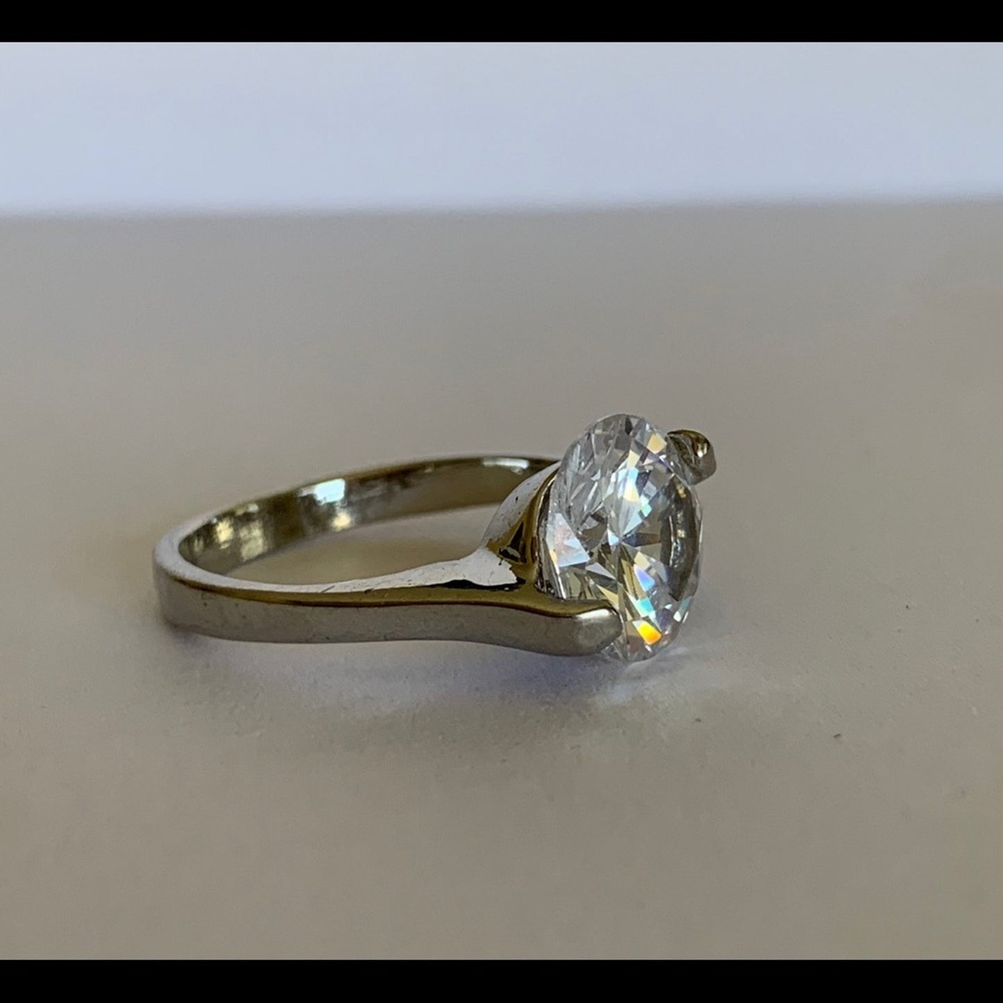 A silver-toned ring with a large, clear cut gemstone in a prong setting, resting on a neutral gray surface.