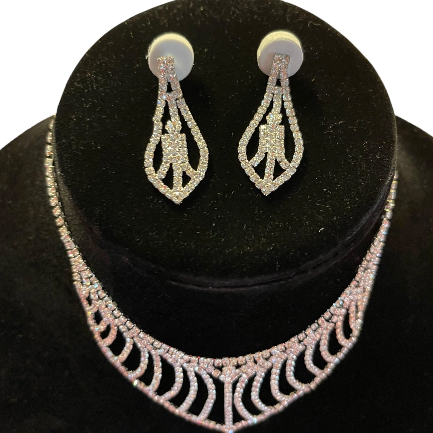 A silver-tone jewelry set featuring a statement collar necklace and matching drop earrings, both embellished with clear rhinestones, displayed on a black velvet jewelry bust.