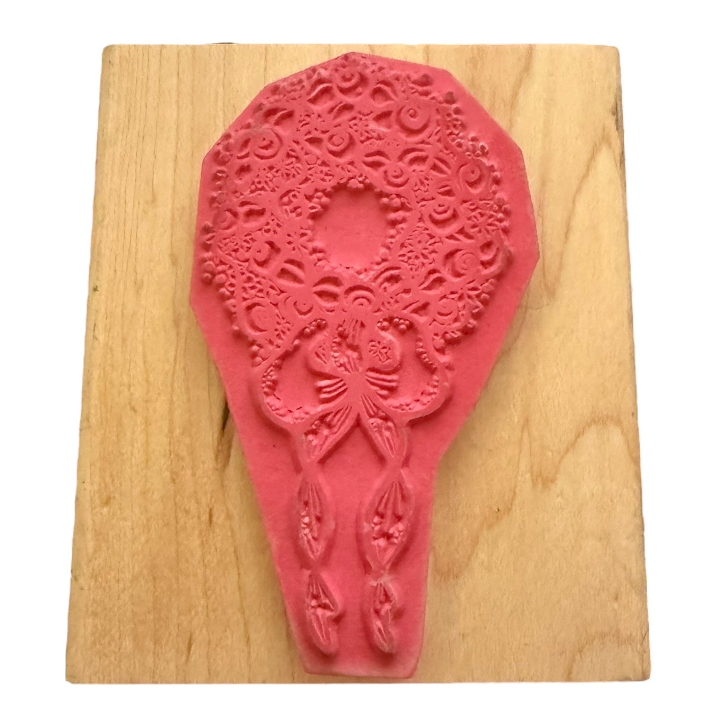 A pink stamp with an intricate floral and bow design, displayed on a light wood block against a white background.