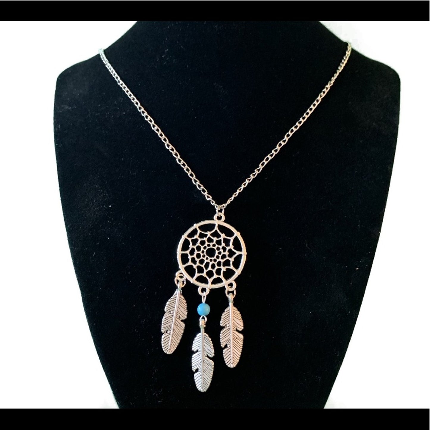 A dreamcatcher-style pendant necklace displayed on a black jewelry bust. The necklace has a silver-tone chain and a circular web design with three hanging feather charms in silver accented by a small turquoise bead.