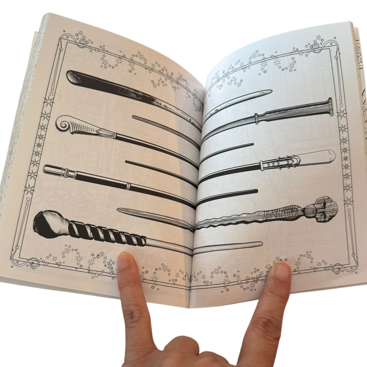 An open coloring book showing black line art illustrations of various fantasy wand designs, held open by two fingers against a white background.
