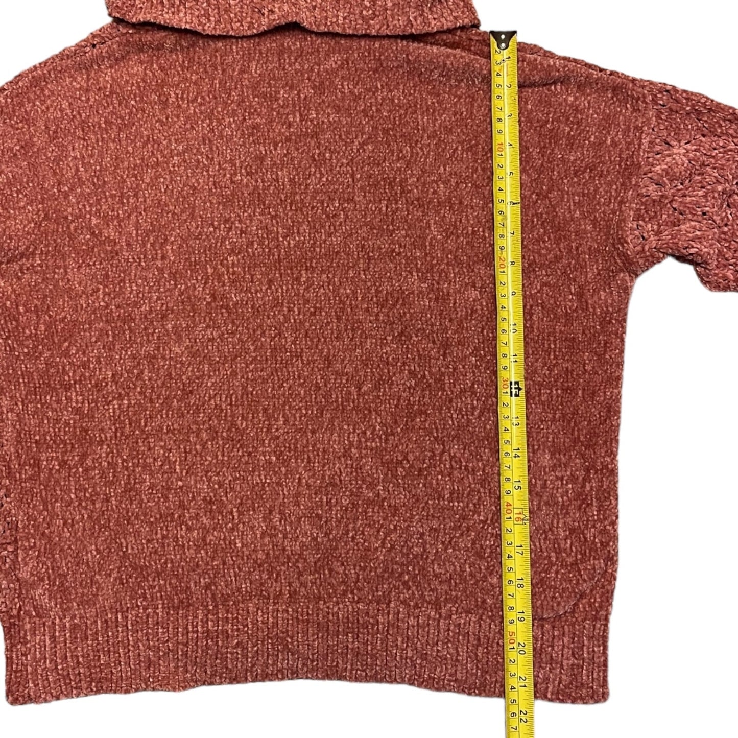A pink colored knit sweater laid flat on a white background, with a yellow tape measure standing vertically along the side to show the garment's length.