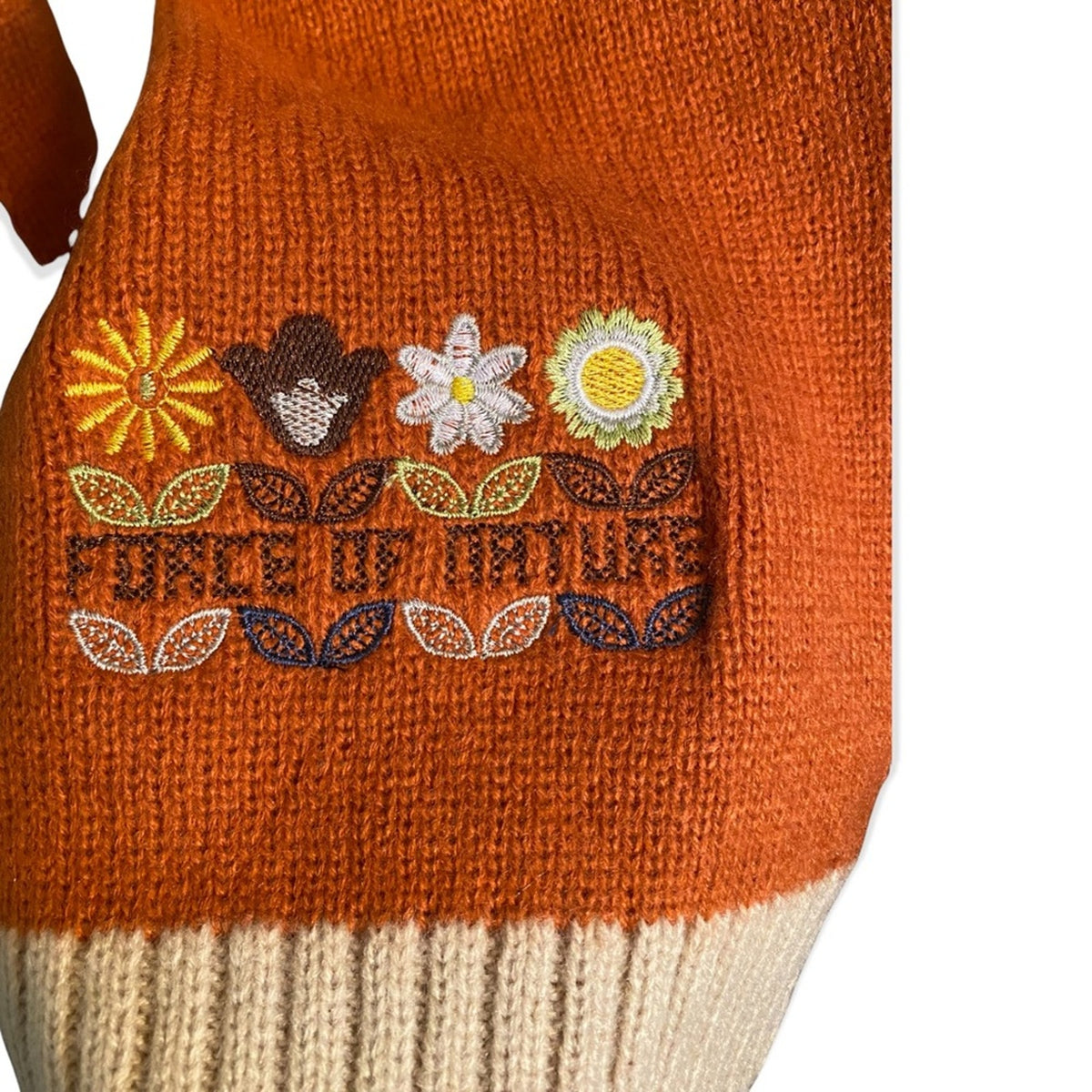 Close-up of an orange knit sleeve with a cream ribbed cuff and embroidered floral motifs and the words “FORCE OF NATURE” on the front.