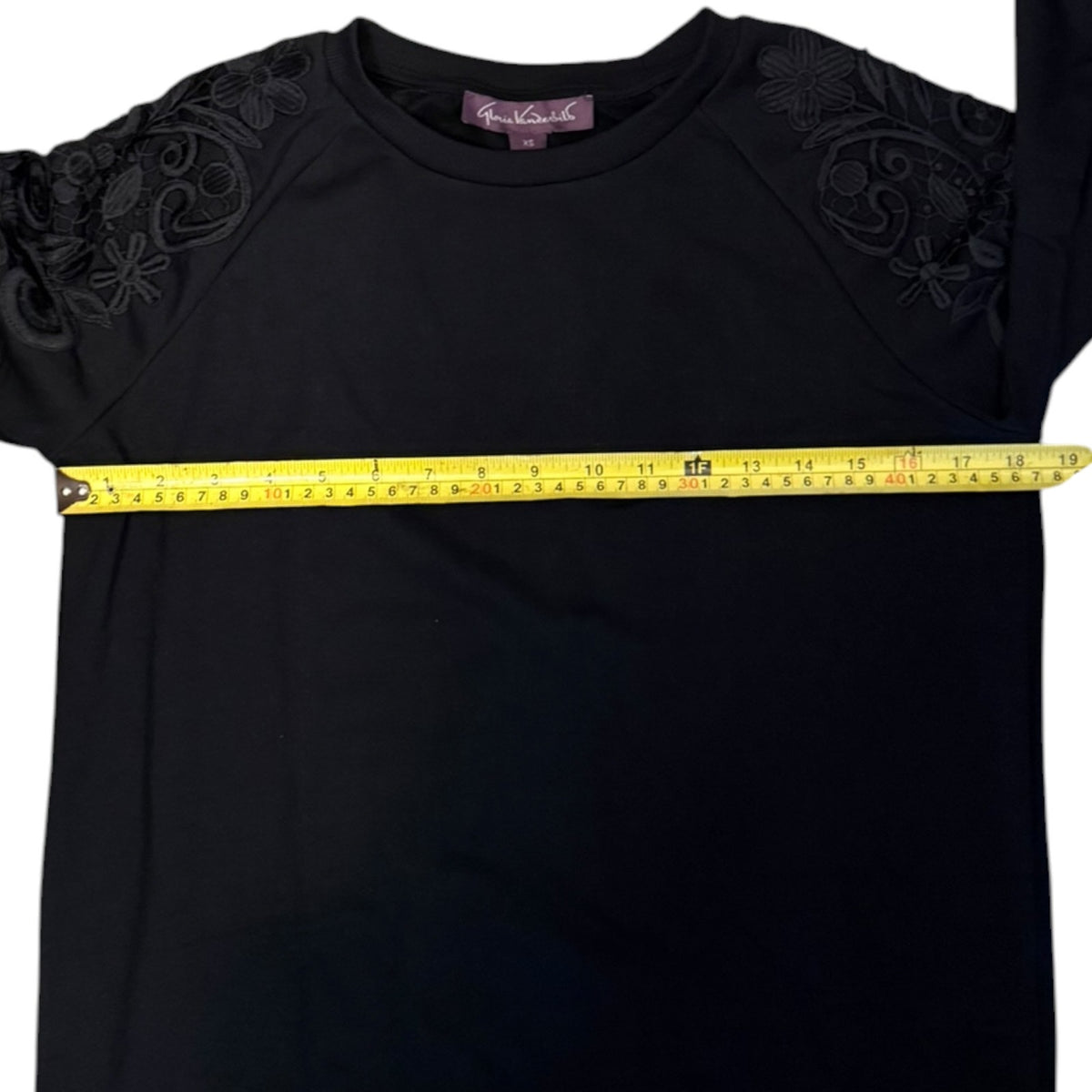 A black long-sleeve sweatshirt with floral lace applique on the shoulders, shown with a yellow tape measure stretched horizontally across the chest to indicate size.