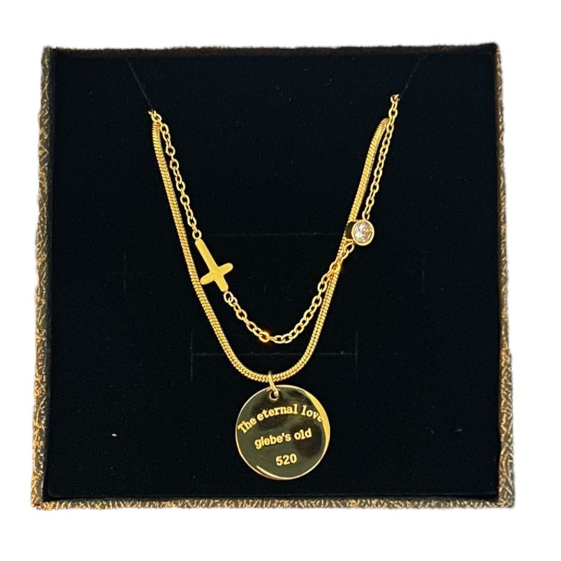 A gold layered necklace displayed on a black velvet box. The necklace features a gold cross pendant, a small round clear stone, and a round gold disc pendant engraved.