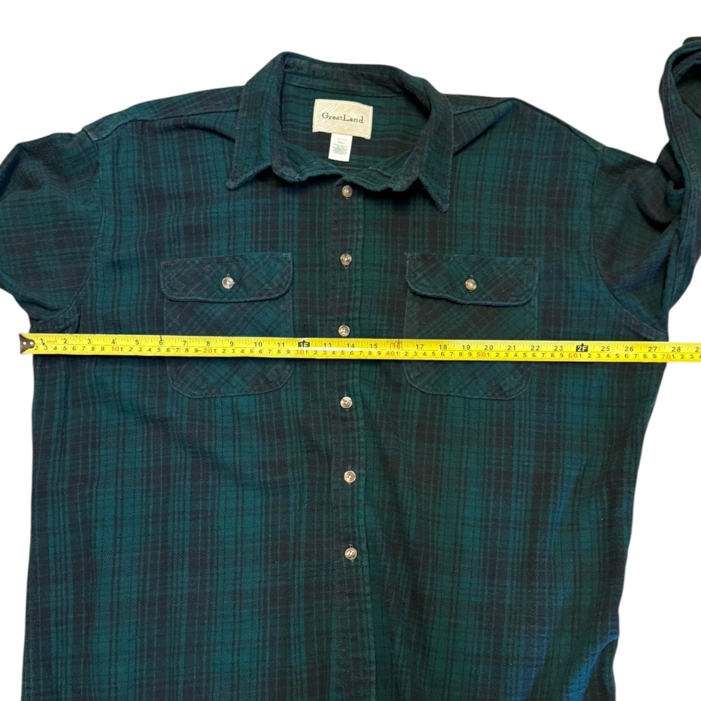 A dark green and black plaid button-up shirt laid flat with a yellow tape measure stretched horizontally across the chest to show the width.