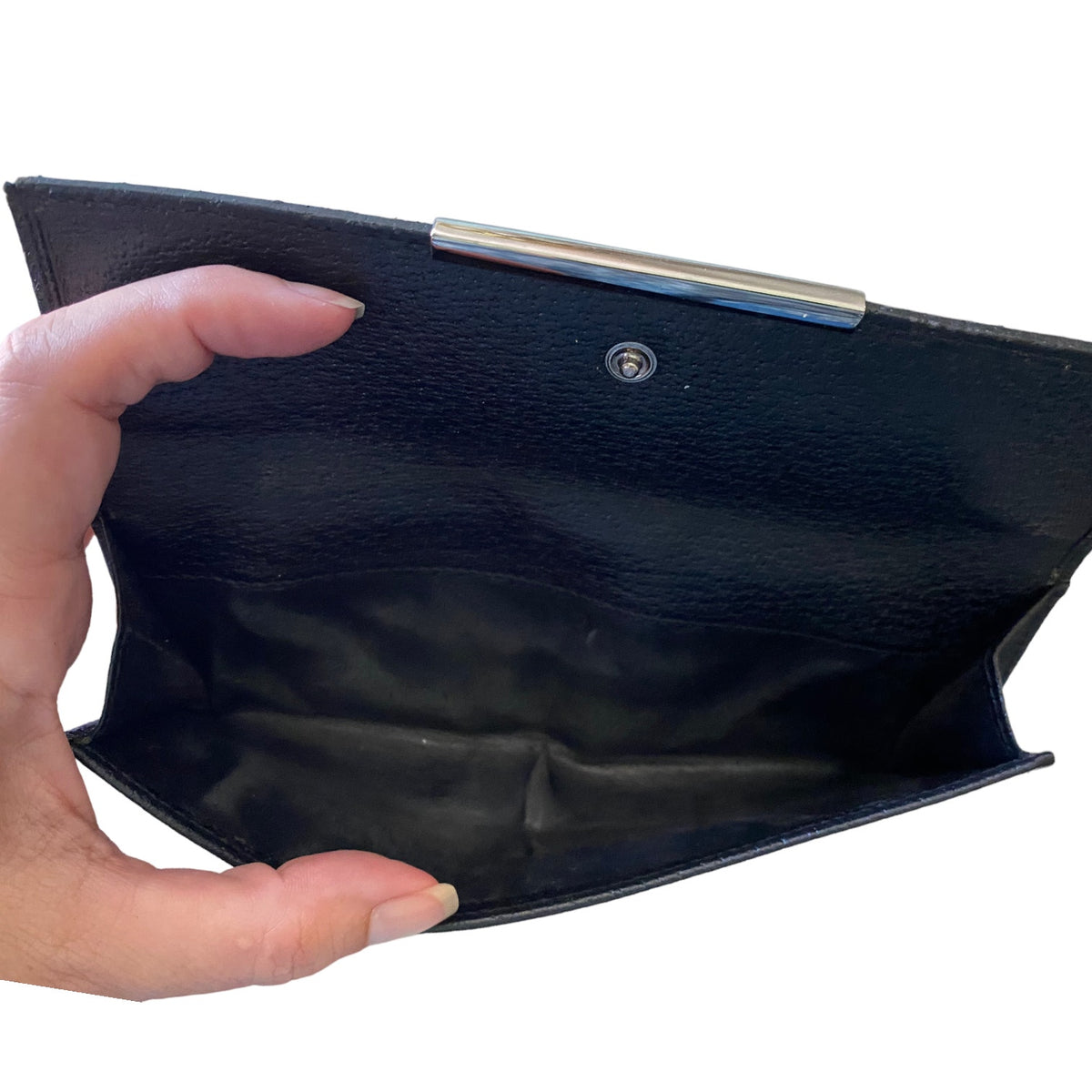 A hand holding open a black textured leather wallet with a silver metal bar clasp, showing the interior lining against a white background.