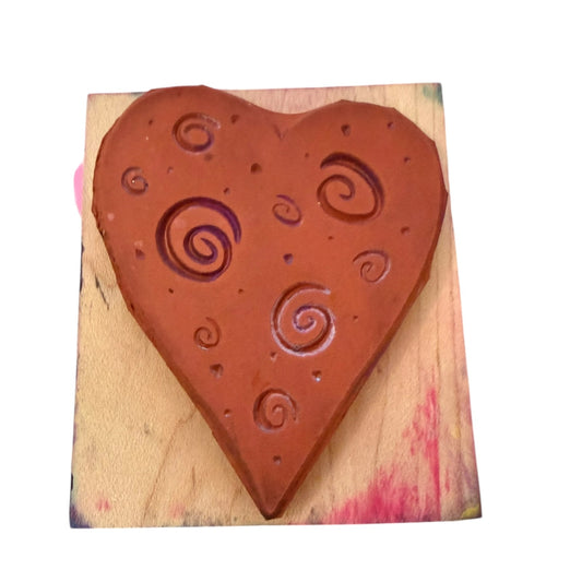 A heart-shaped stamp with embossed swirl patterns, mounted on a square wooden block with some ink splatters, isolated on a white background.