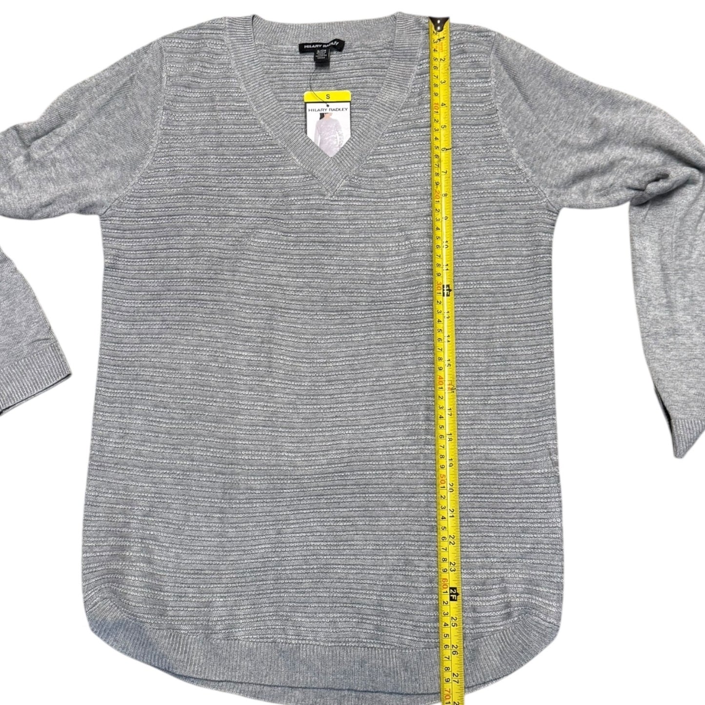 A gray knit sweater with a V-neck and long sleeves, laid flat on a white background with a yellow measuring tape running vertically down the center to show the garment's length.