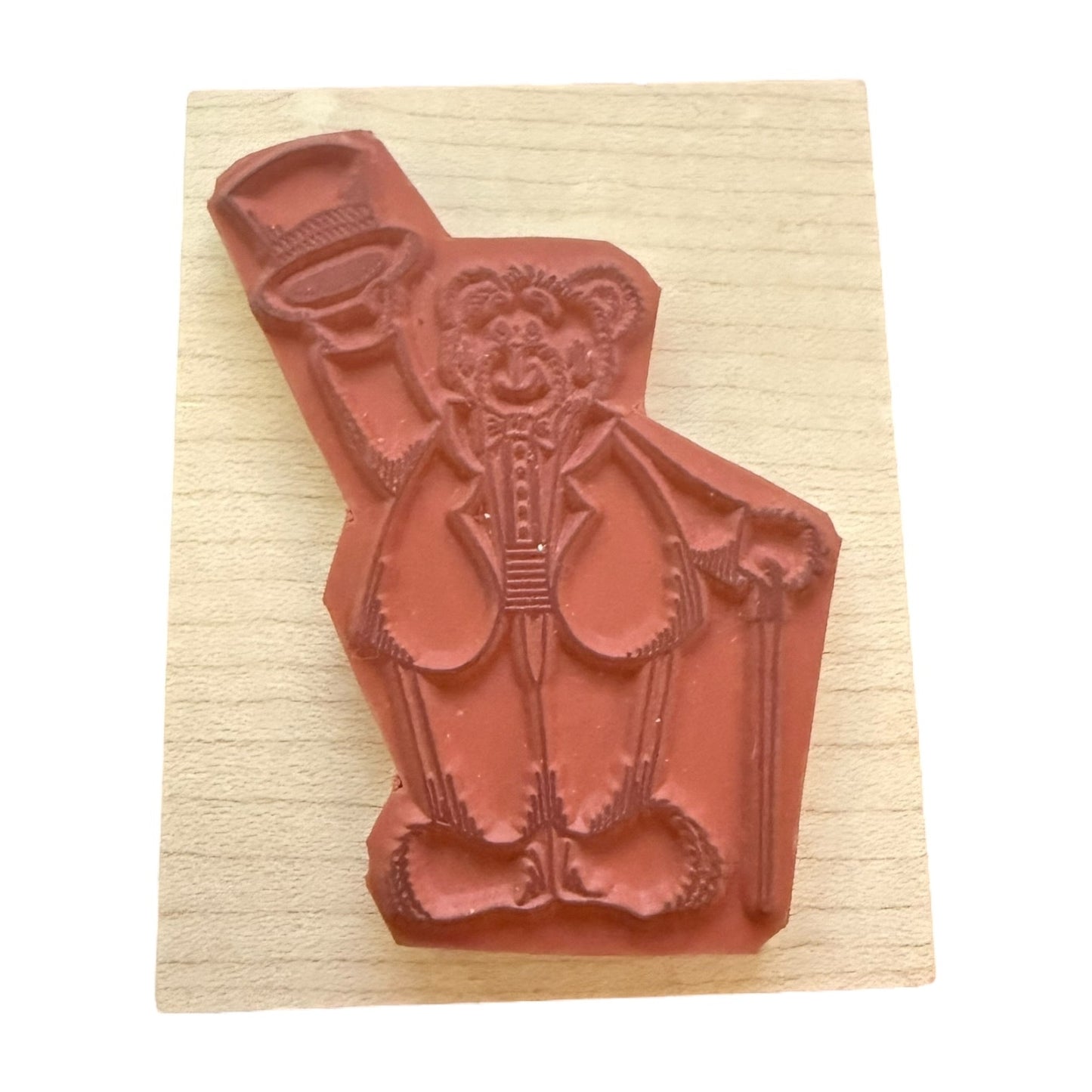 A terracotta-colored rubber stamp featuring a teddy bear character in a suit holding a cane, mounted on a rectangular light wood block against a white background.