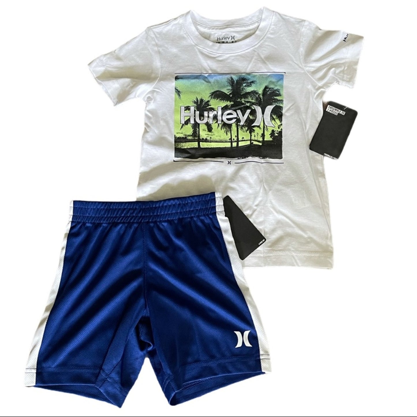 A two-piece boys outfit set featuring a white Hurley t-shirt with a colorful palm tree graphic and blue athletic shorts with white side stripes, laid flat on a white background with black tags attached.