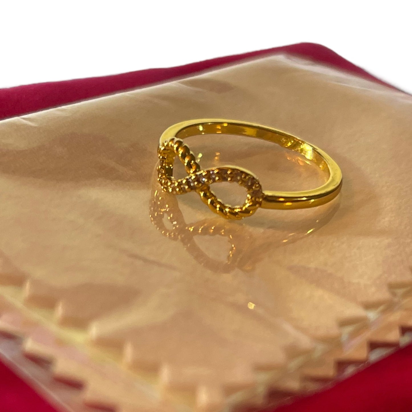 A gold infinity ring with small clear stones, displayed on a clear plastic sleeve over a red velvet jewelry box.