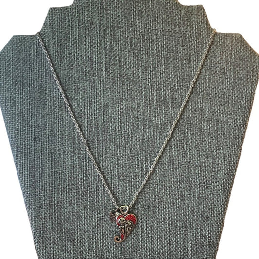A silver-toned necklace with a small heart-shaped pendant featuring red and gold details, displayed on a fabric jewelry bust against a white background.