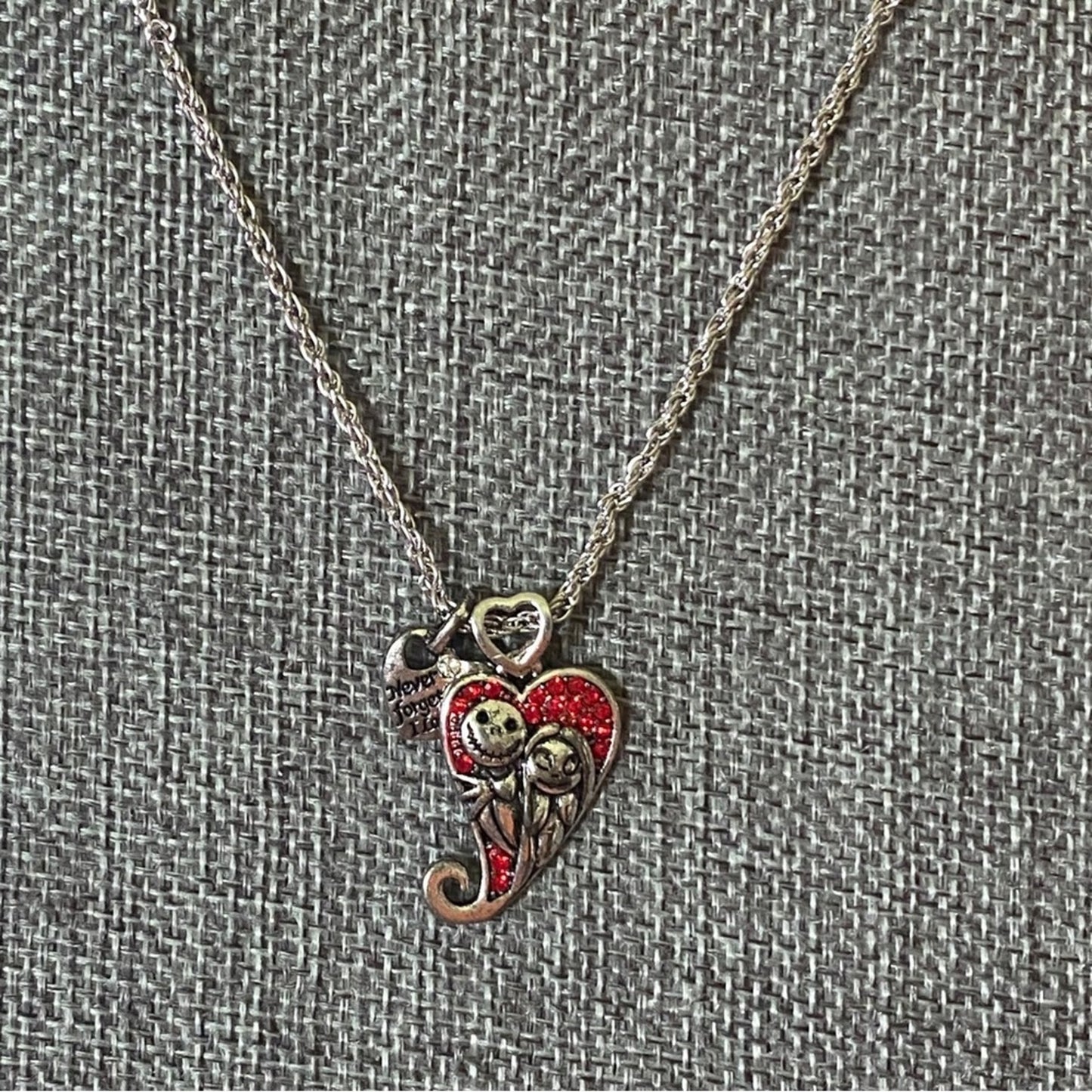 A silver-toned necklace with a heart-shaped pendant featuring a Jack Skellington and Sally design, accented with red glitter and a small heart charm, displayed on a textured gray fabric background.