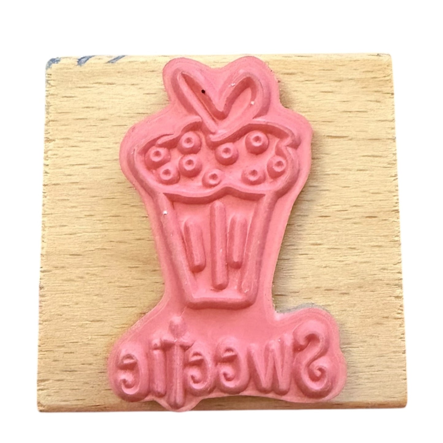 A pink rubber stamp featuring a cupcake design with the word 'Sweetie' at the bottom, mounted on a square wooden block.
