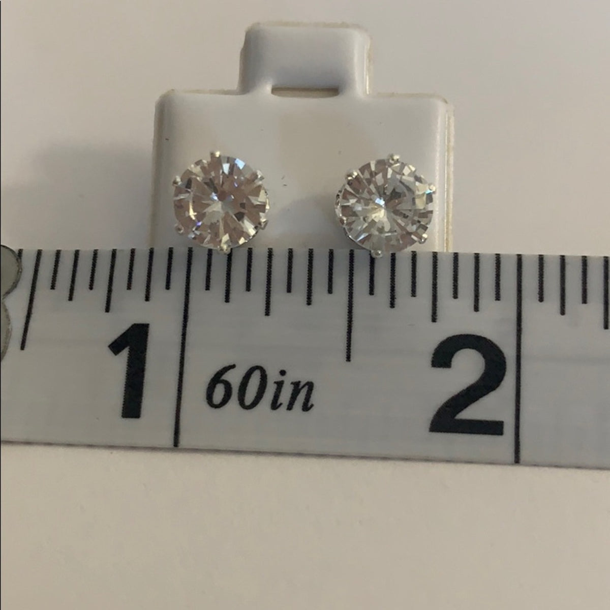 A pair of round-cut stud earrings displayed on a white plastic card above a measuring ruler to show size. The earrings appear clear and reflective, set against a plain light background.