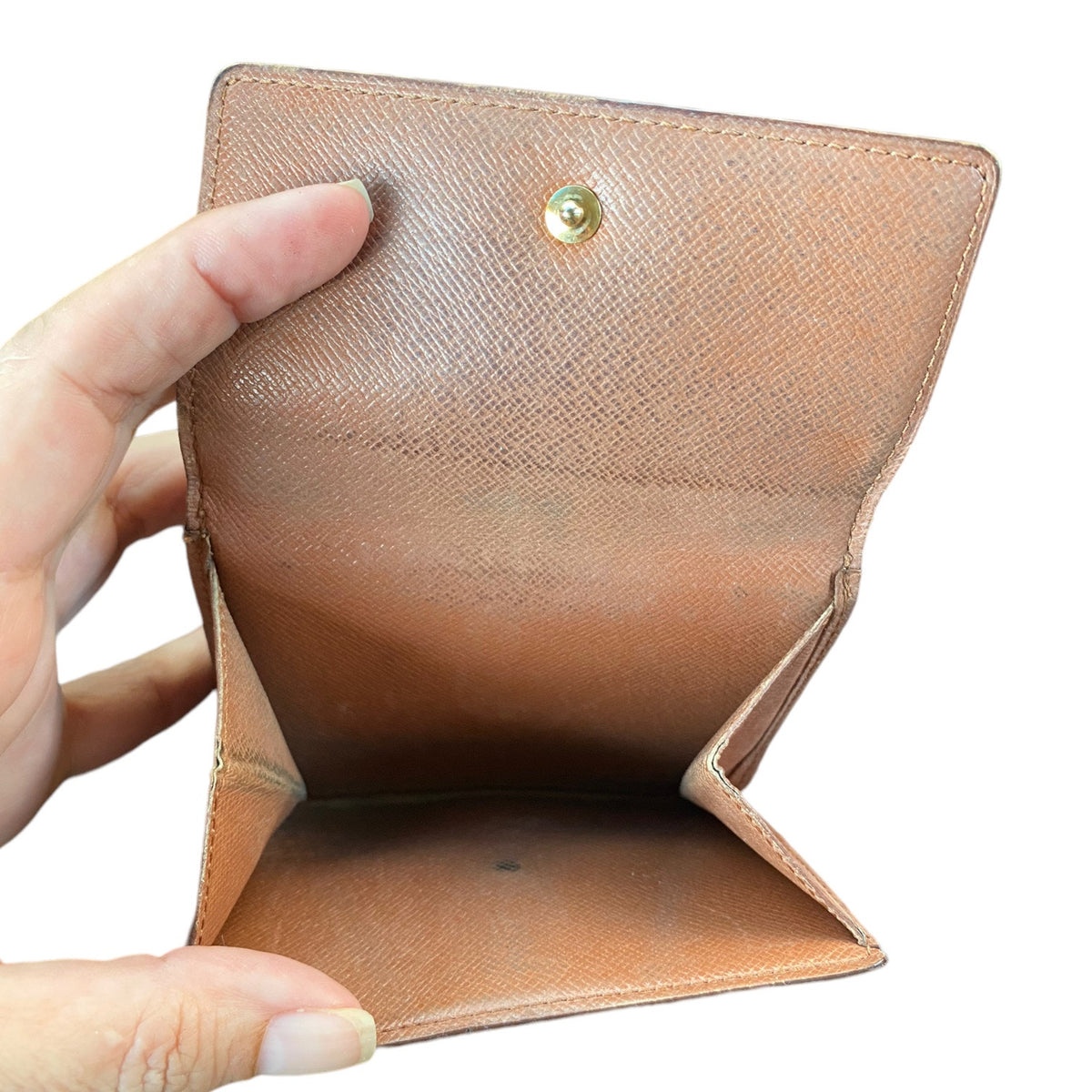 A tan textured wallet held open by a hand, showing its interior compartments and a gold-tone snap button closure, set against a plain white background.