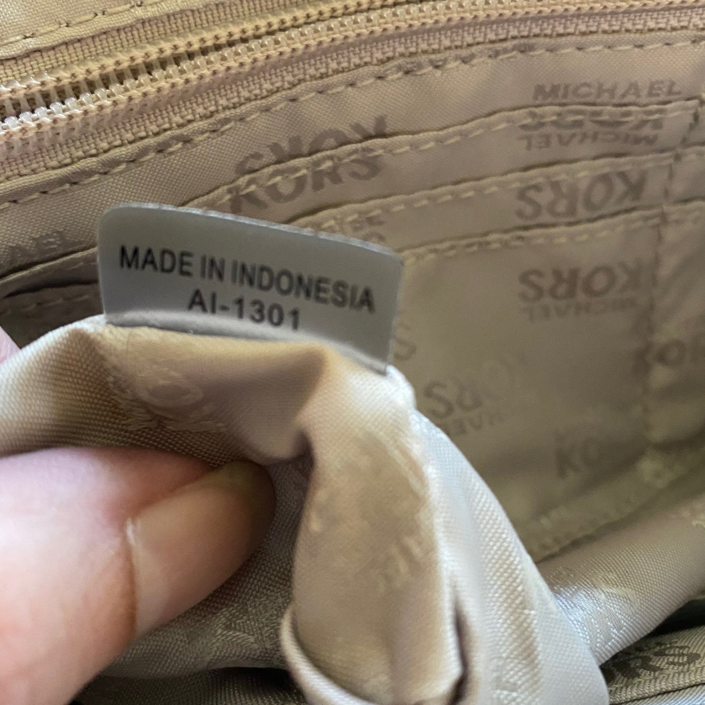 Close-up of a beige Michael Kors interior label reading 'MADE IN INDONESIA AI-1301' held by a finger, with the brand name repeated on the lining.