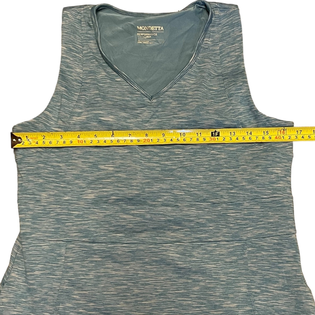 A sleeveless V-neck dress in a heathered blue color, shown with a yellow measuring tape stretched horizontally across the chest to indicate the waist or torso measurement.