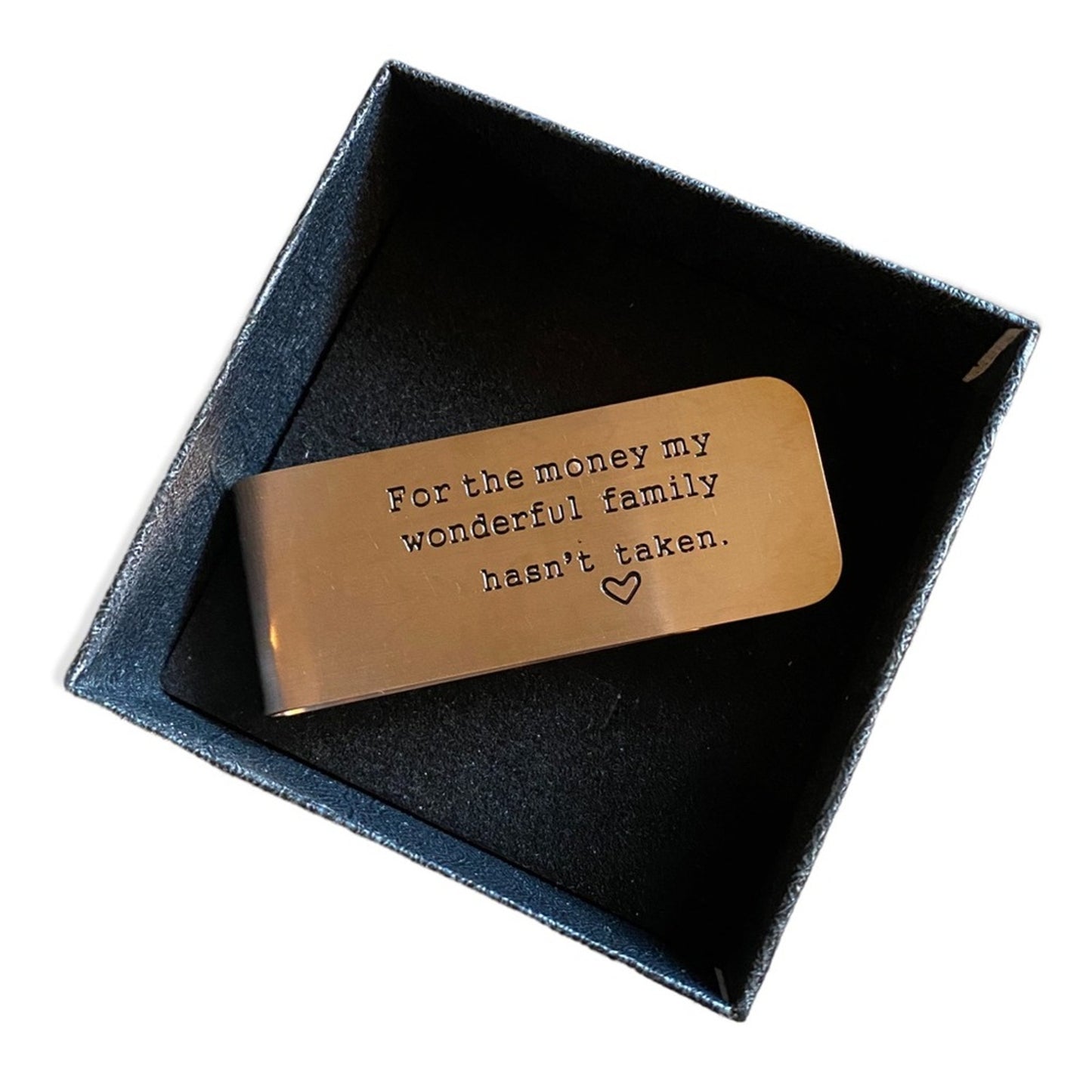 A rectangular money clip engraved with the phrase 'For the money my wonderful family hasn't taken.' and a small heart, presented inside a dark blue-gray gift box on a white background.