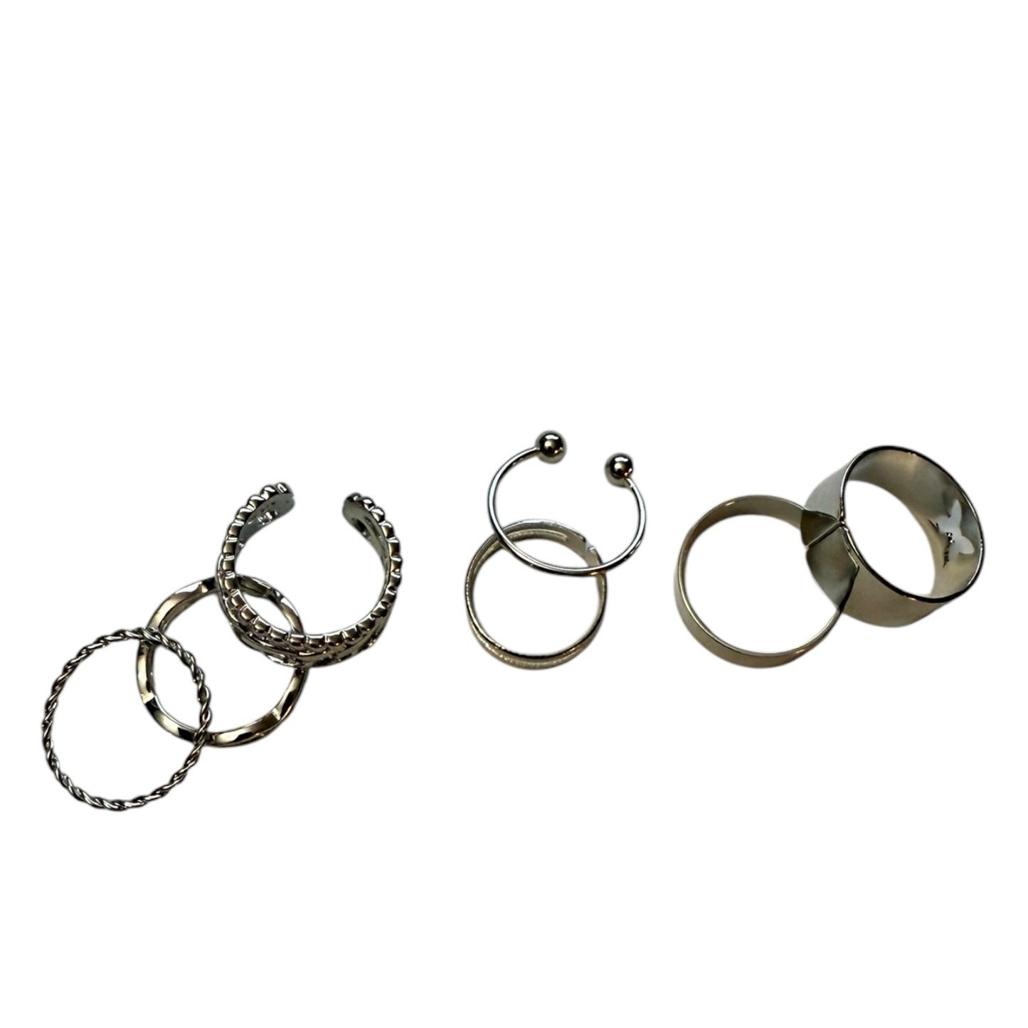 Seven silver-toned metal rings and cuffs displayed on a plain white background, including textured open cuffs, a small hoop with a ball end, and two smooth bands.