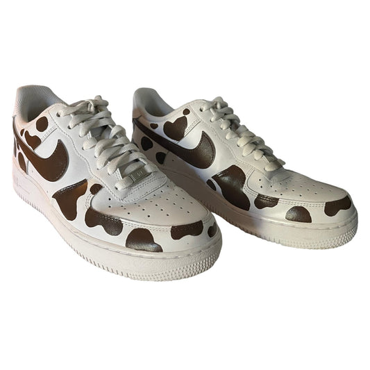A pair of white Nike Air Force 1 Low sneakers featuring a brown cow-print pattern and a brown Swoosh logo.
