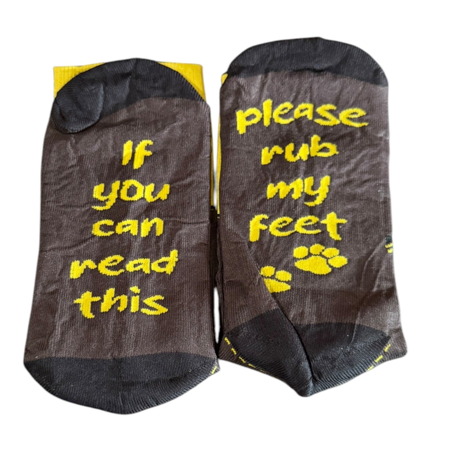 A pair of dark brown crew socks with yellow text reading 'If you can read this' on one sock and 'please rub my feet' with paw prints on the other, shown against a plain white background.