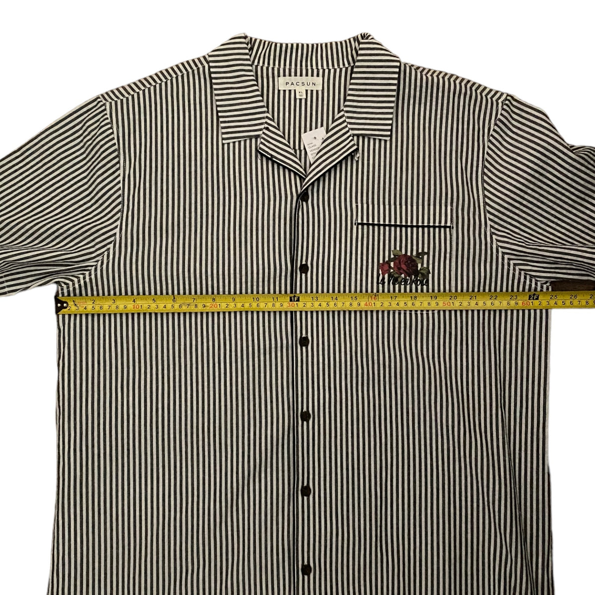 A short-sleeve button-up shirt with thin black and white vertical stripes, featuring a small embroidered rose on the left chest. A yellow tape measure is laid horizontally across the chest to indicate the garment's width.