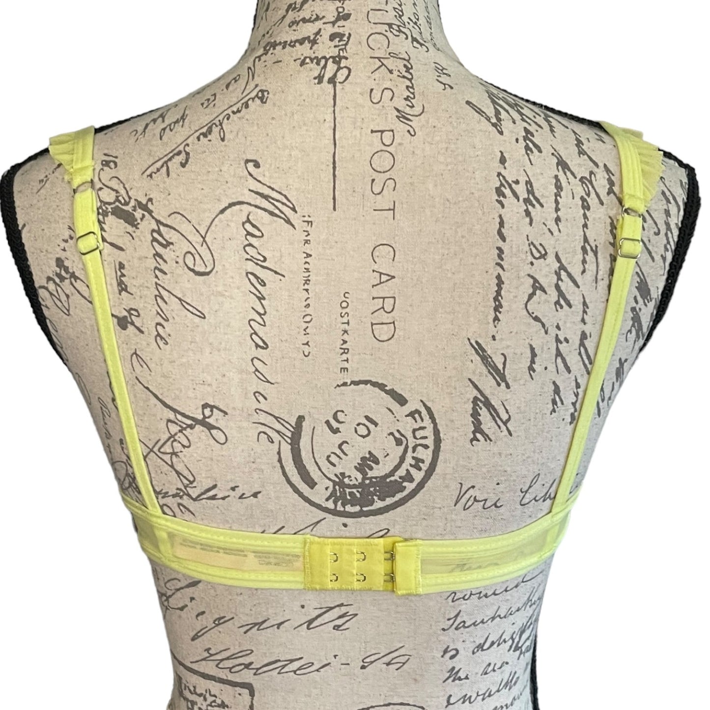 A neon yellow strappy bra displayed on a vintage-style fabric mannequin torso with a cursive script print.