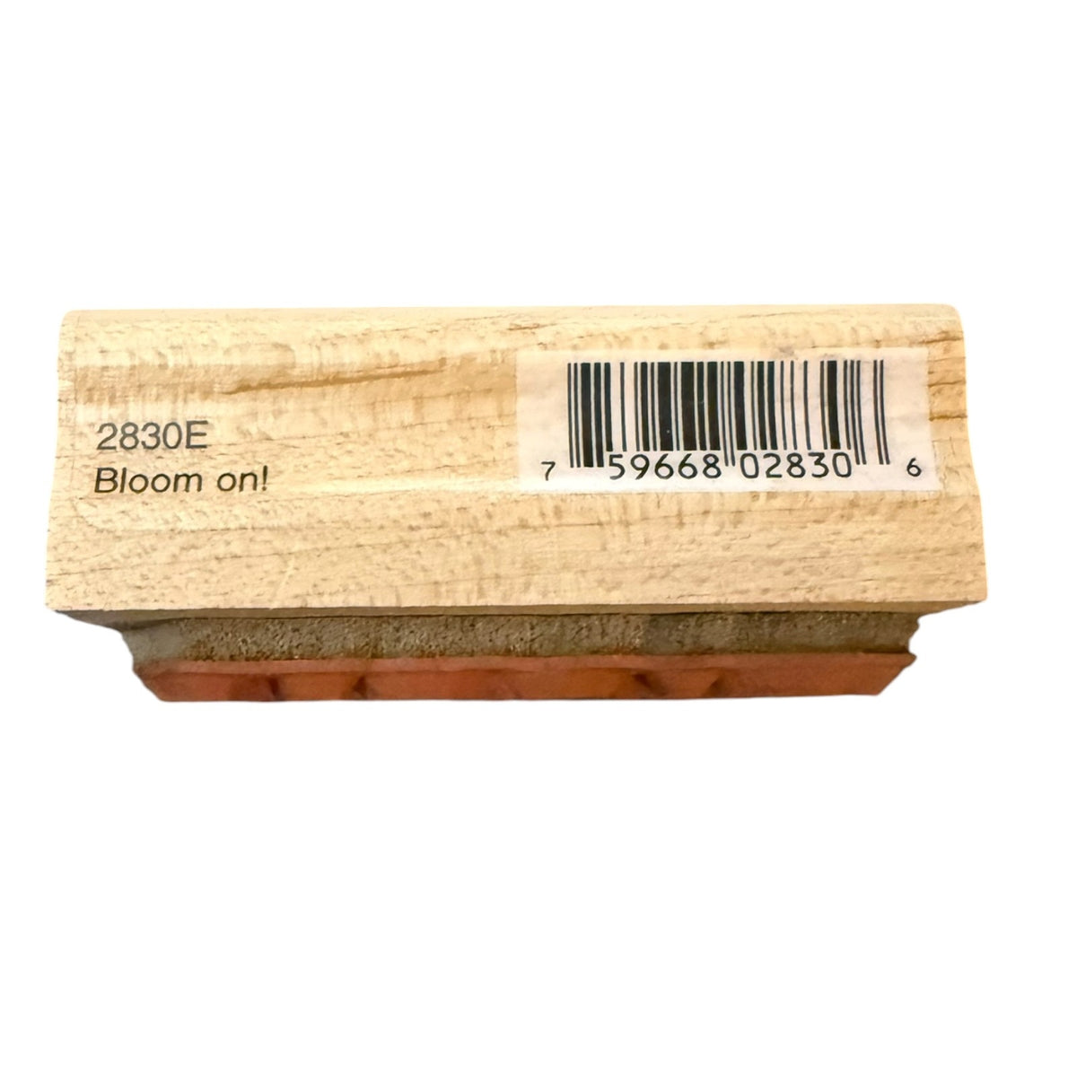 A rectangular wooden stamp block with a light natural wood grain surface, printed with the text '2830E Bloom on!' on the left and a barcode with numbers on the right. The bottom edge shows a reddish-brown rubber stamp face.