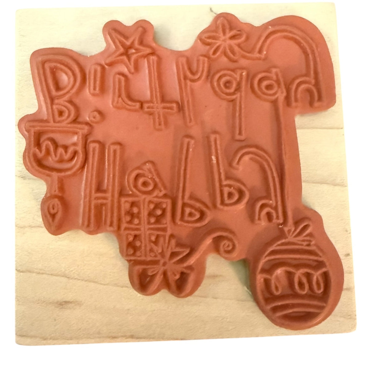 A terracotta-colored rubber stamp featuring birthday-themed letters in stylized lettering, mounted on a light-colored wooden block.