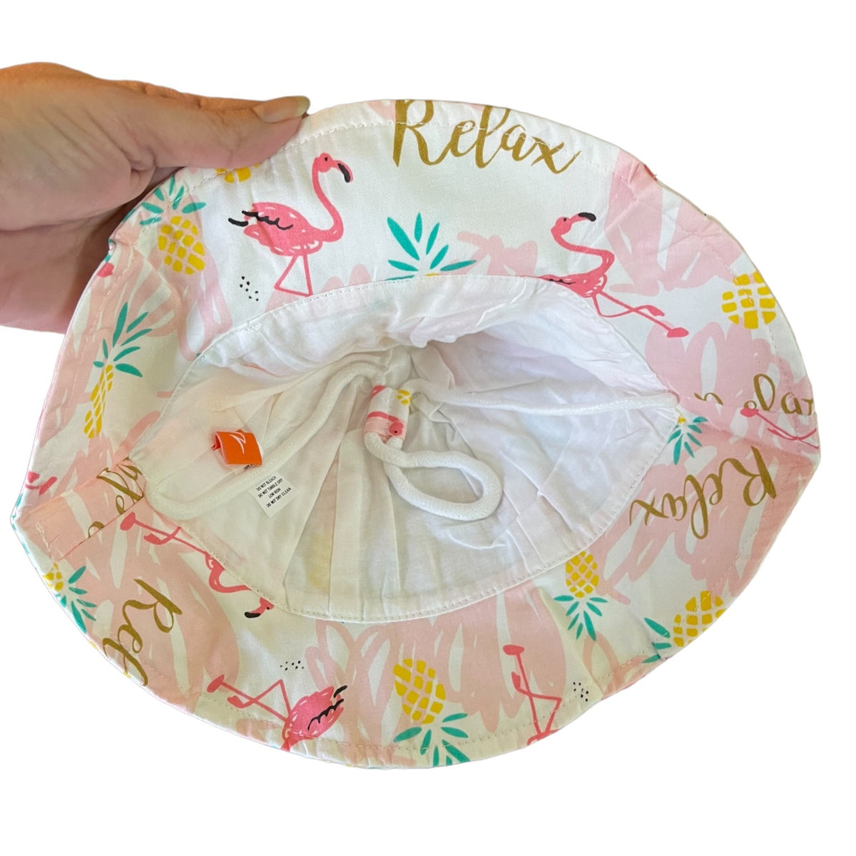A hand is holding a round sun hat against a white background. The hat features a light pink and white tropical print with pink flamingos, pineapples, and gold lettering that reads 'Relax'.