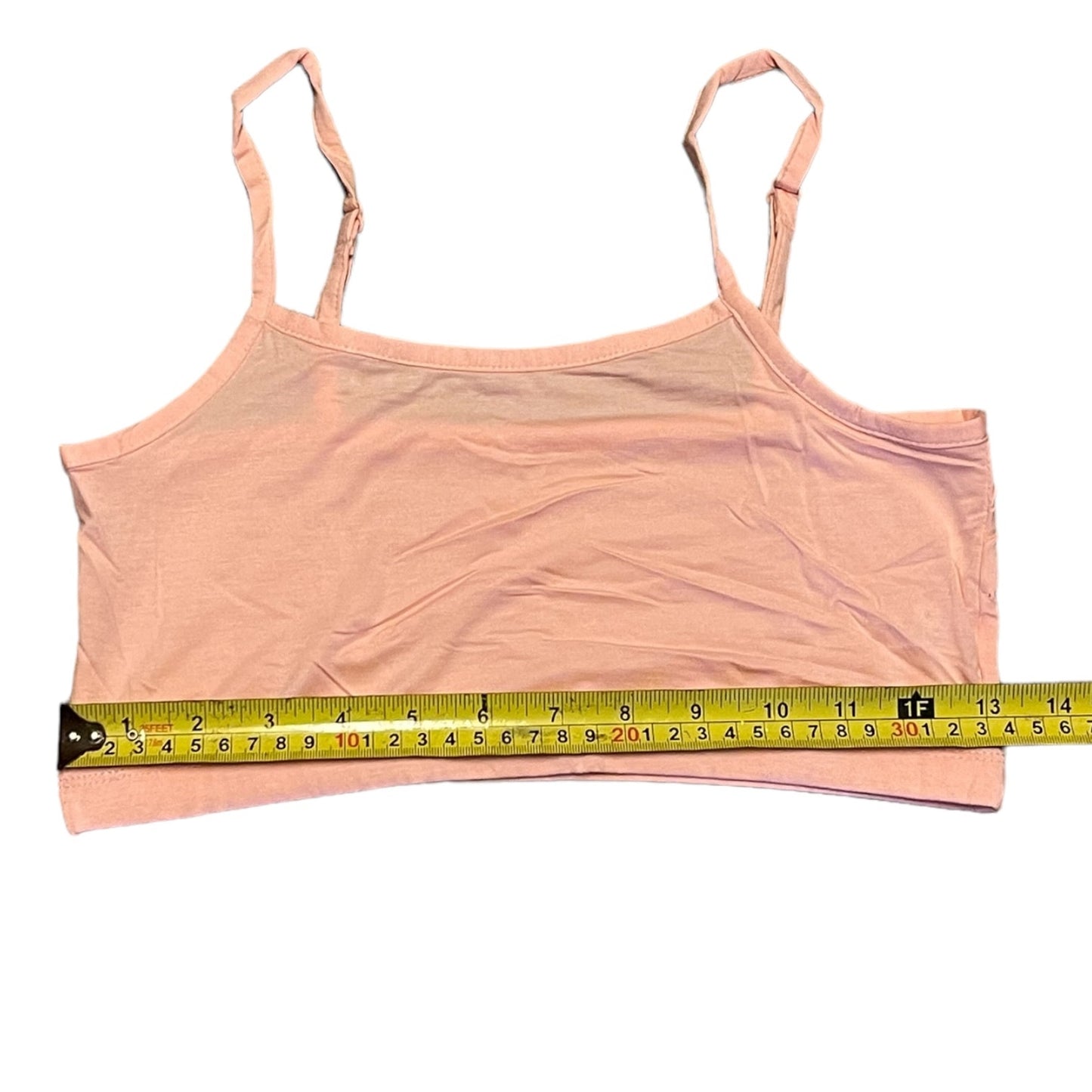 A light pink spaghetti-strap crop tank top laid flat on a white background, with a yellow tape measure stretched horizontally across the front to show the width.