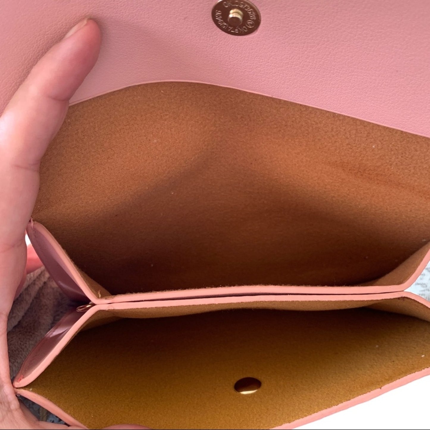 Close-up of a pink leather purse shown open, with a hand holding the left side to display the interior. The inside is lined in brown suede-like material, and a gold-tone snap button is visible at the top.