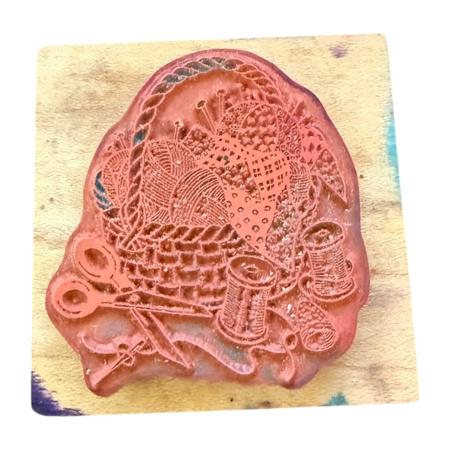 A rubber stamp featuring a detailed design of a basket, thread spools, scissors, and needles, resting on a wooden block.