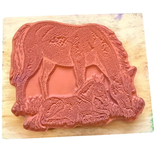 A terracotta-colored rubber stamp featuring an embossed design of a mare and foal, displayed on a light wood block against a white background.